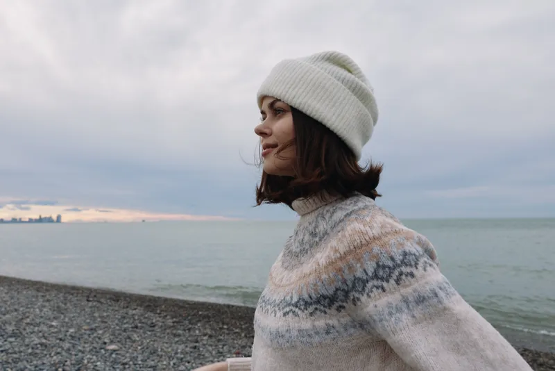 Woman in a knitted sweater and beanie on a pebbled beach with the ocean backdrop and a cloudy sky, capturing a calm outdoor moment