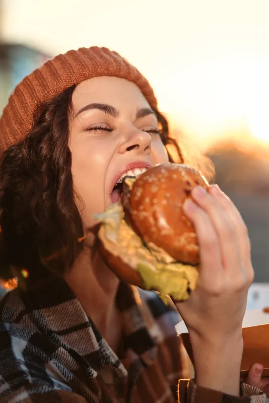 Woman biting a burger outdoors in a beanie, candid smile and juicy bite at golden hour glow, authenticity and mindful living vibe, emotional storytelling of urban cozy food moment.