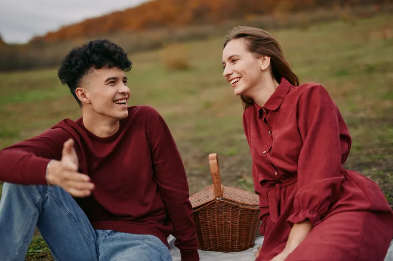 A young couple sitting on a picnic blanket in a grassy field, sharing a genuine conversation and laughter; an authentic candid moment capturing natural connection and relaxed emotions.