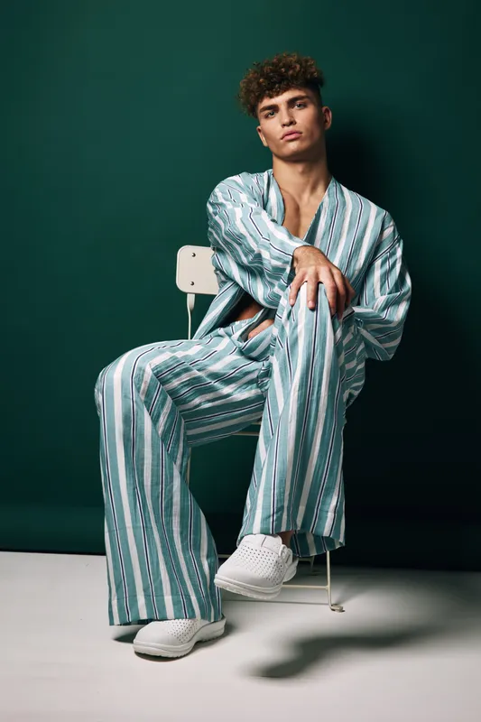 Young man sitting on chair wearing striped loose pants and shirt with white sneakers against dark green background in studio portrait.