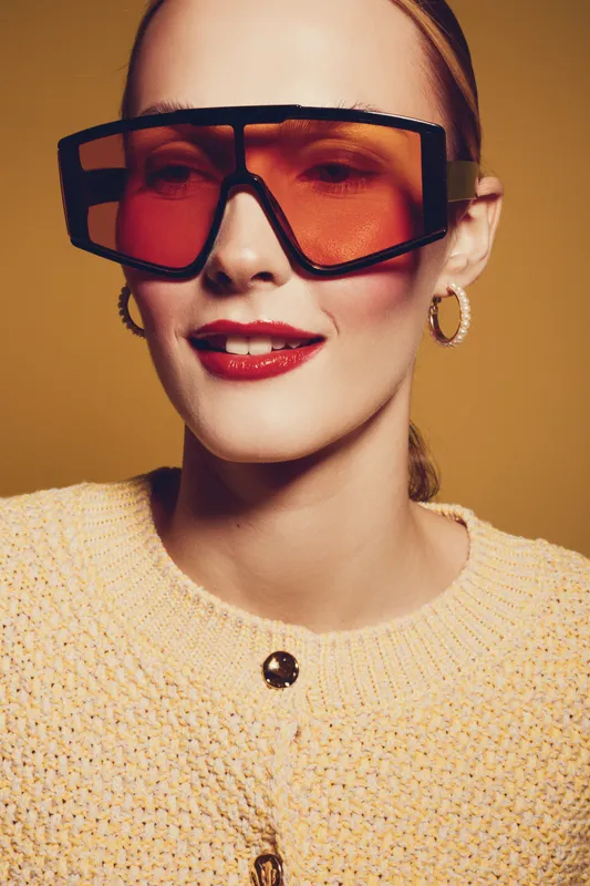 Stylish woman with oversized orange sunglasses and gold hoop earrings wears knitted beige sweater. Close-up portrait on warm brown background. Concept of fashion, accessories, and modern style.