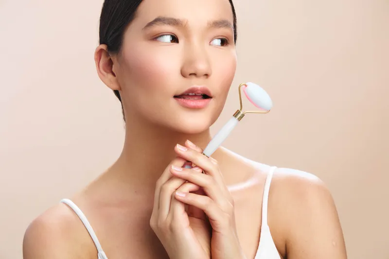 asian woman holds a facial roller near her face, showcasing a skincare routine in a soft, minimal setting. she gazes calmly, radiating gentle care, creating a peaceful beauty moment and a focus on