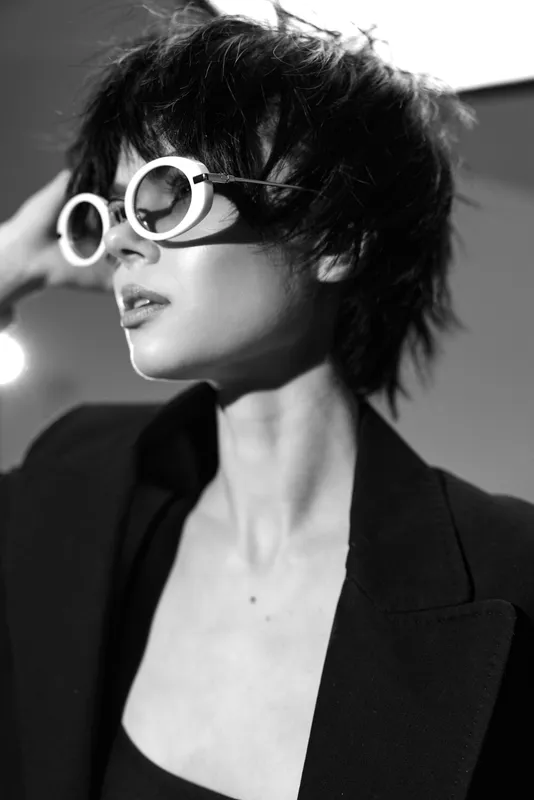 Black and white portrait of a stylish woman wearing round sunglasses and a dark jacket, expressing confidence and sophistication in a modern setting.