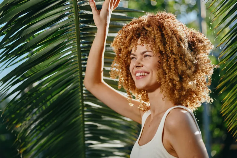 smiling woman with curly hair enjoying sunny day outdoors, surrounded by green palm leaves and natural light, wearing white top and radiating happiness