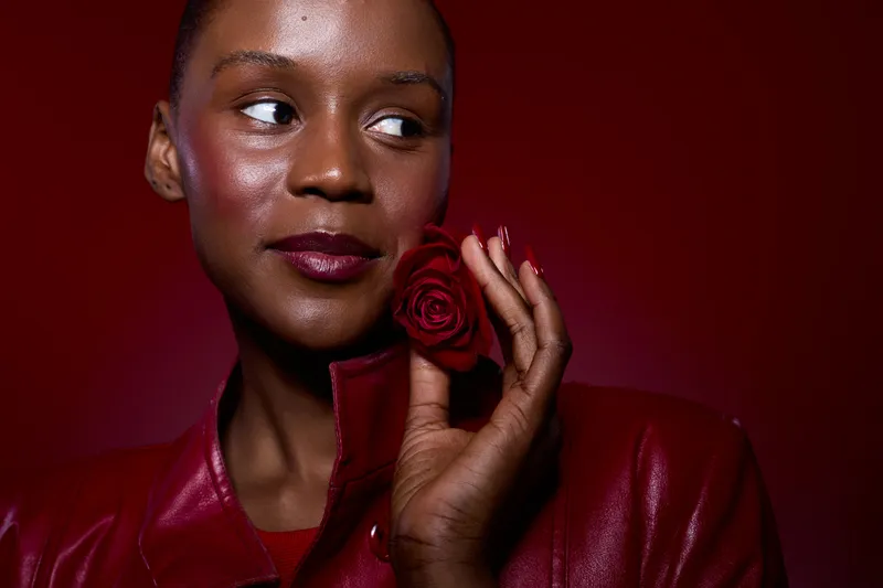 Close-up of a beautiful black woman with dark skin holding a red rose near her face. She wears a red leather jacket and looks to the side against a red background, concept of elegance and beauty.