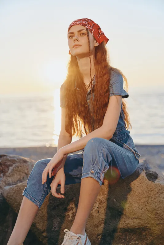 Lifestyle woman in modern boho-western denim street style sits on rocks by the sea with warm film color tones, relaxed pose, and natural sunlight at sunset.