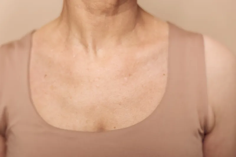 Closeup of middle aged woman chest and neck with natural skin texture and wrinkles wearing beige top. Concept of aging, skin care, and natural beauty on neutral background.