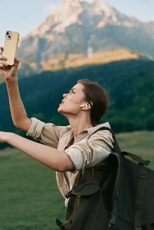 Smiling woman with backpack taking a selfie outdoors in front of a dramatic mountain landscape. Casual hiking scene, travel lifestyle, and outdoor exploration with soft natural light.