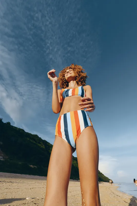 Young woman with curly red hair enjoys a sunny day on the beach wearing a colorful striped high-waisted bikini under a partly cloudy sky.