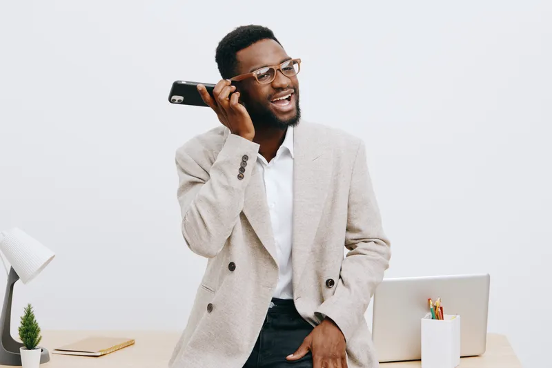 Smiling man talking on smartphone indoors, wearing beige blazer and glasses, standing at desk with laptop and stationery, professional and joyful expression, modern office environment, lifestyle conce
