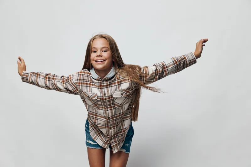 Girl smiling with arms outstretched wearing plaid shirt and denim shorts against white background. Happy child plays and poses for casual portrait in studio environment.