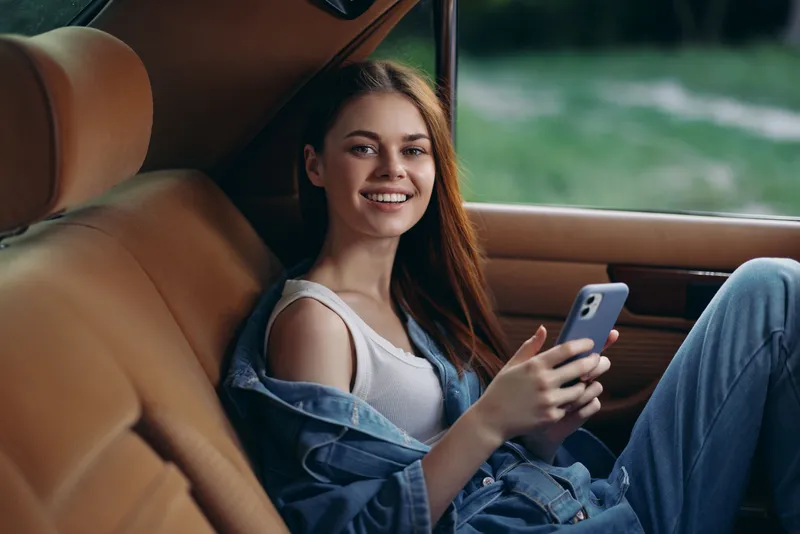 Young woman with long hair smiling while using a smartphone in a car, wearing casual denim clothing and relaxed outfit. Emphasizes modern lifestyle and leisure.