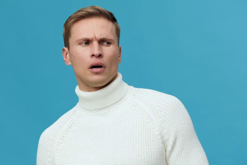 Man with confused expression wearing white turtleneck sweater isolated on blue background studio portrait. People facial expression concept
