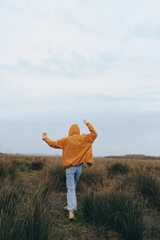 Woman lifestyle, outdoor field, jumping, orange jacket, blue jeans, tall grass, cloudy sky, countryside horizon, feeling freedom and joy, soft natural light, calm wellness