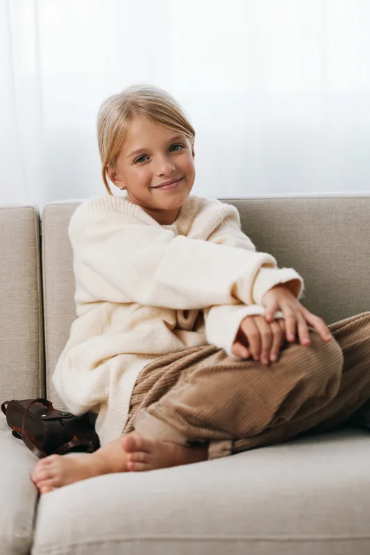 Young girl sits comfortably on a light sofa, wrapped in a soft cream sweater, barefoot and relaxed, smiling softly at the camera as natural daylight fills a calm living room.