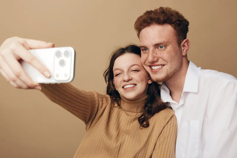 Young couple taking a selfie with a smartphone. They smile and pose closely together against a plain beige background. Concept of friendship, technology, and casual lifestyle.