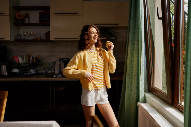 Young woman in a yellow cardigan and striped top enjoys a sunny morning indoors beside a kitchen window, radiating a warm calm and casual cheer.