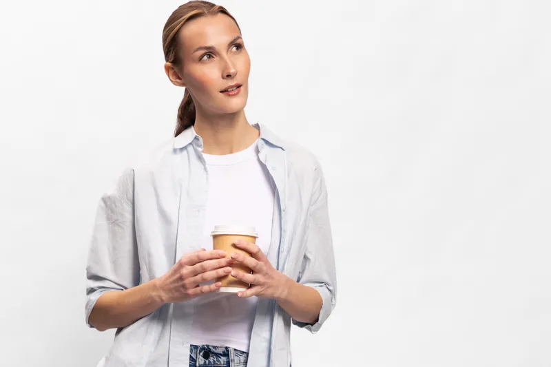 Young woman with a ponytail holding a paper cup looks thoughtfully aside. Casual outfit on white background. Concept of relaxation, break, and casual lifestyle.