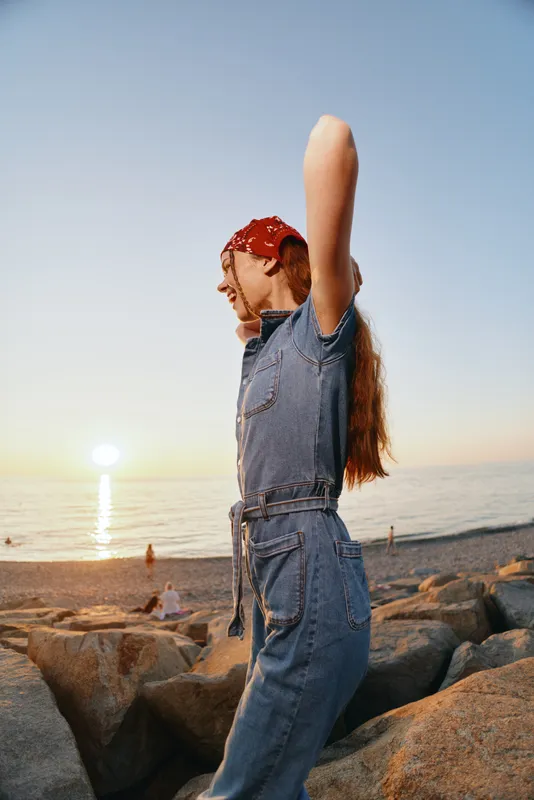Lifestyle woman in modern boho-western denim street style enjoying sunset by the rocky beach with film color warmth and relaxed carefree mood.