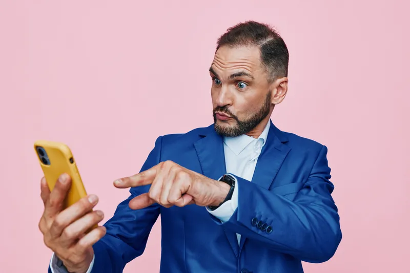 A business professional man against a solid pink backdrop, focused on his smartphone, posture alert, and pointing gesture that conveys curiosity, confidence, and engaged emotion.