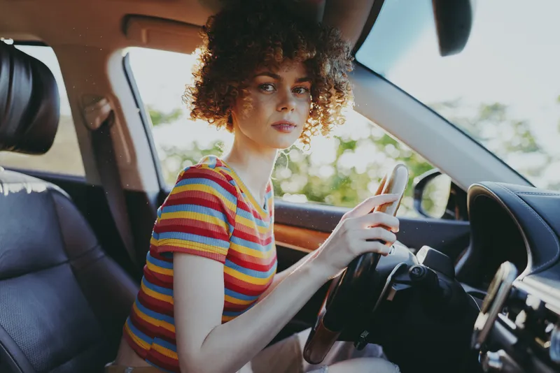Woman driver sits in a car, lifestyle portrait with retro film color, casual striped shirt, curly hair, daylight glow, focused expression, urban street mood, daytime commute vibe, candid style