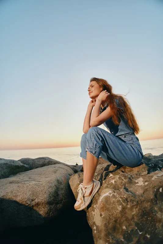 lifestyle woman in modern boho-western denim street style sits thoughtfully on rocks by the sea, captured with warm film color tones in sunset light