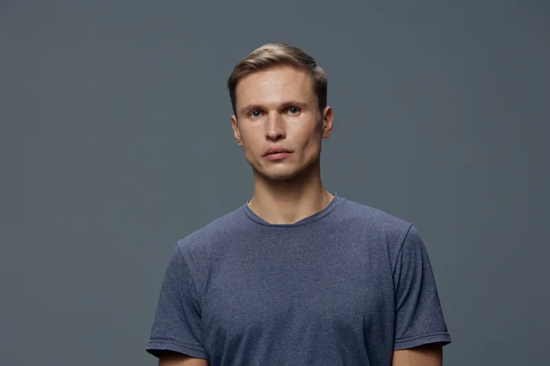 Full body portrait of a serious young Caucasian man with short light brown hair, wearing a casual blue T-shirt, looking directly at camera, isolated on a plain gray background, studio shot. People con