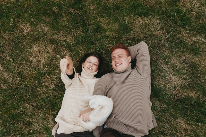 Couple lying on grass in cozy sweaters, smiling and relaxed, with a small dog nearby. Lifestyle moment outdoors, showcasing connection, comfort, and happiness in a natural setting.