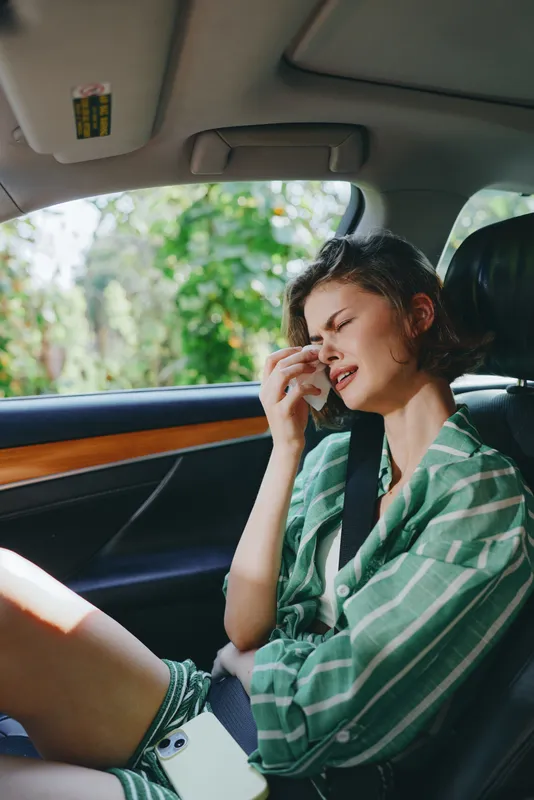 A woman in a green striped shirt sits inside a car, rubbing her eye and showing a tired, overwhelmed expression.