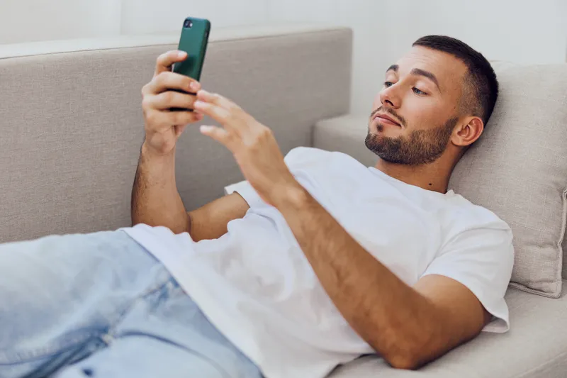 Relaxed man checking phone while lounging on sofa, showcasing soft masculinity and the blend of personal and work life in a modern, comfortable setting.