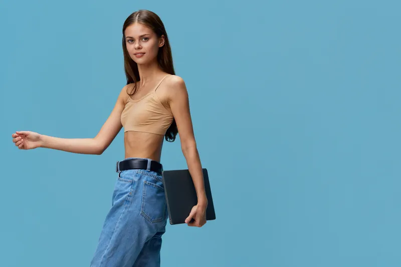slim woman in beige top and blue jeans walking with laptop against plain blue background, casual young adult, technology and style concept