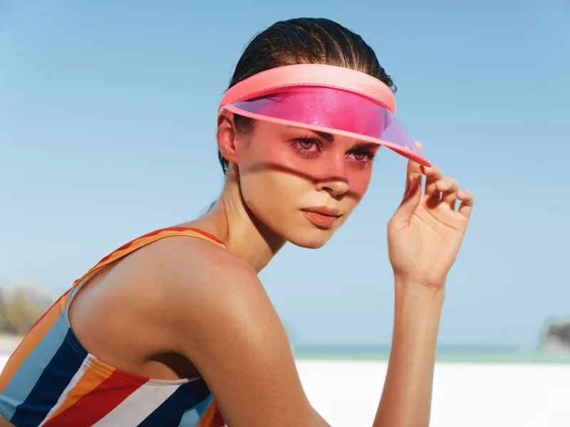 A close view of a young woman on a sunny beach wearing a pink visor and striped swimsuit, her gaze forward as bright light highlights sunlit skin and a vibrant mood.