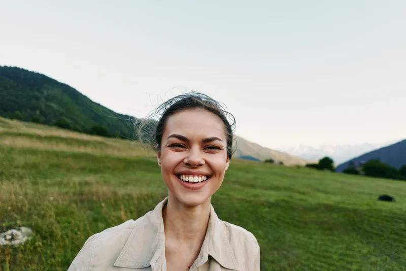 Young brunette woman smiles warmly in a sunlit meadow with distant mountains and a clear blue sky, conveying an outdoorsy, carefree lifestyle.