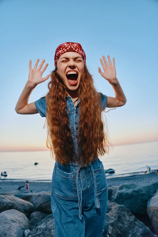 Lifestyle woman in modern boho-western denim outfit, expressing joy and energy on the beach during sunset in film color style with casual street vibe and long hair.