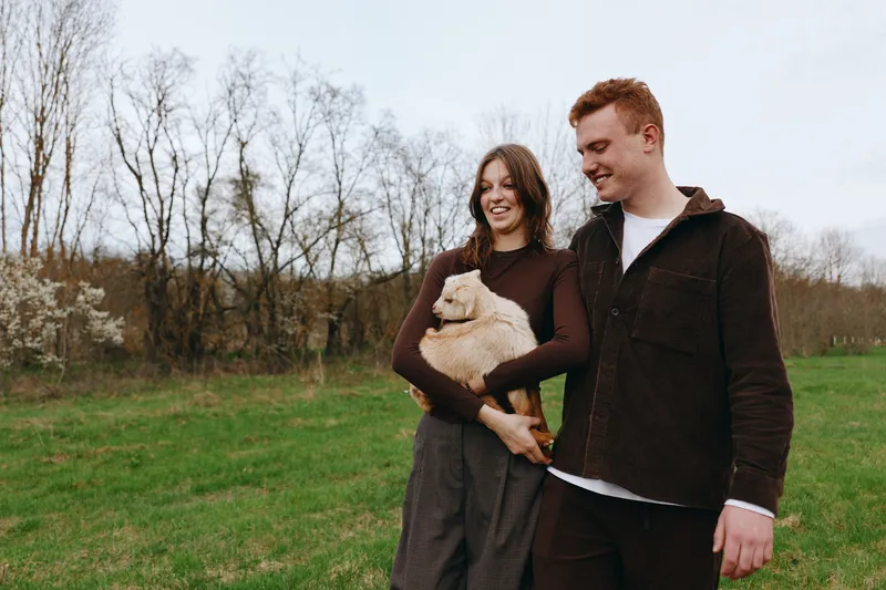 Outdoors lifestyle couple with a gentle goat kid, smiling, sunlit breeze, relaxed posture, natural colors, lush grass, candid moment, warm connection, simple joy, countryside scene