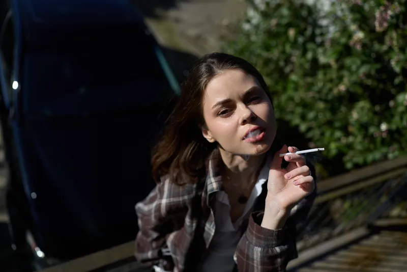 Woman lifestyle outdoor scene with casual mood, smoking a cigarette, wearing plaid jacket, wind in hair, focused expression, urban setting beside a car, candid street portrait, daytime light