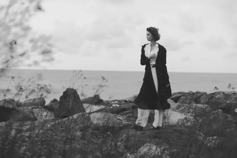 Woman, sea, coast, rocks, coat, fashion, outdoor, monochrome, mood, horizon, a windswept figure on jagged shore stands in a long coat against rugged terrain and open water, creating timeless