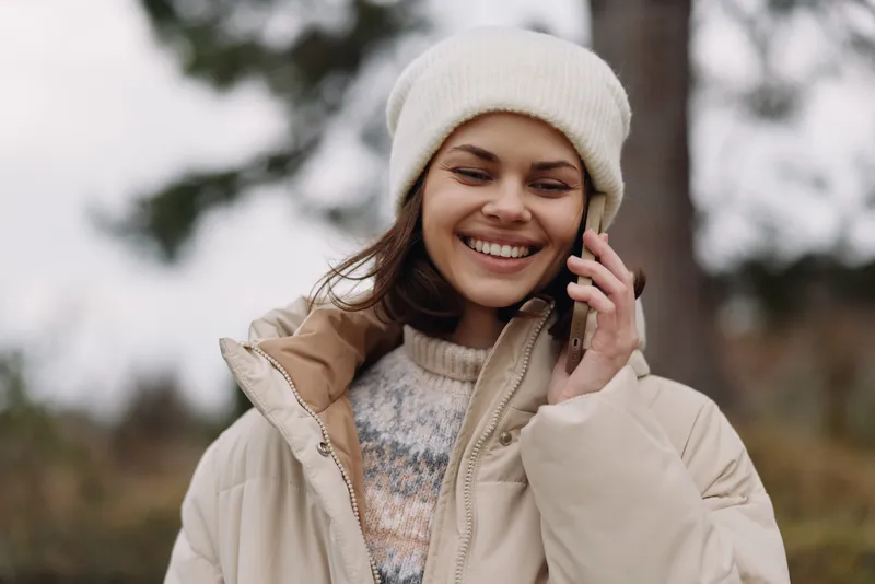 Smiling woman outdoor portrait with phone call in a warm coat and beanie, capturing casual fashion and cozy clothing in natural daylight, an candid lifestyle moment amid outdoor scenery