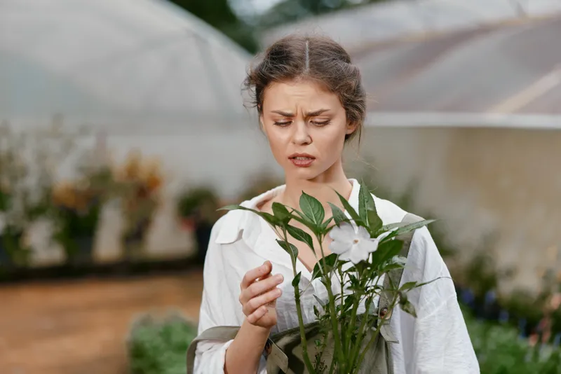 Thoughtful young woman examining flowers in a greenhouse, showcasing emotions of curiosity and care, dressed in a simple white outfit, amidst greenery and soft natural light