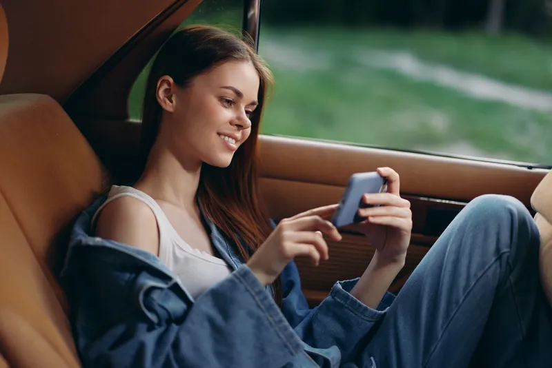 Young woman enjoying her smartphone in a stylish car interior, showcasing casual fashion, happiness, and a relaxed atmosphere on a sunny day.