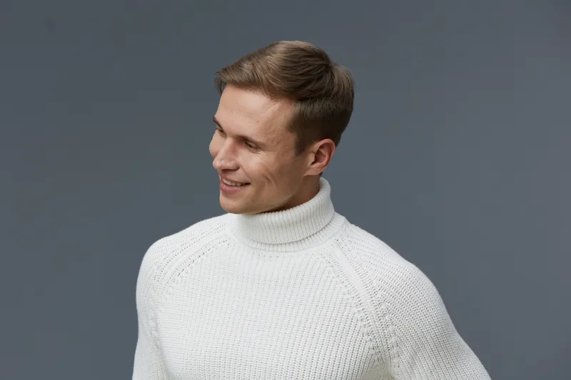 Smiling man dressed in white knitted sweater looking to the side against gray background, portrait of cheerful young male with short brown hair, casual style, positive expression, studio shot. People 