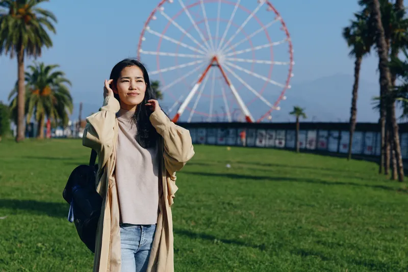 Woman in beige coat standing in sunny park with a bag, distant ferris wheel in background, casual travel mood, open space, leafy trees, blue sky, hopeful vibe, lifestyle shot for outdoors and leisure