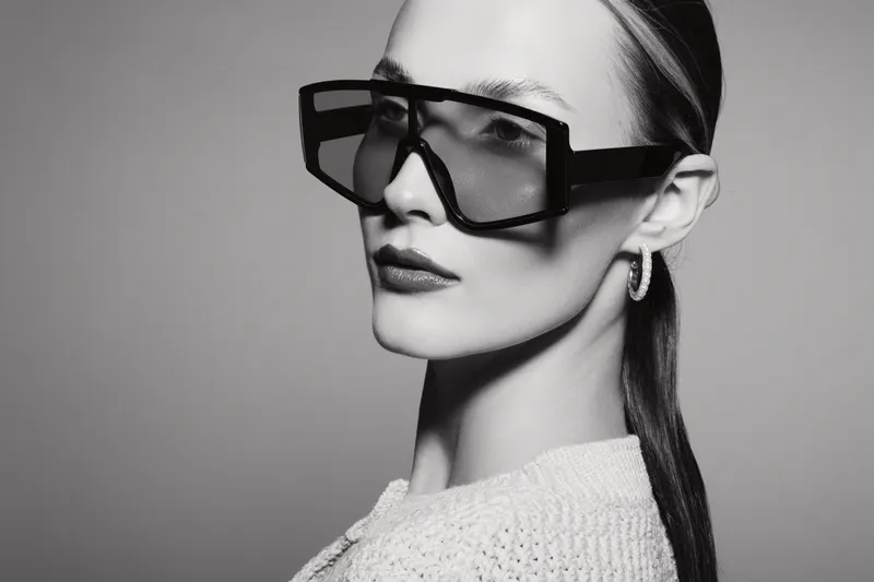 Close-up of a young Caucasian woman with pale skin wearing oversized black sunglasses and textured white clothing. Studio portrait in black and white highlights fashion and elegance.
