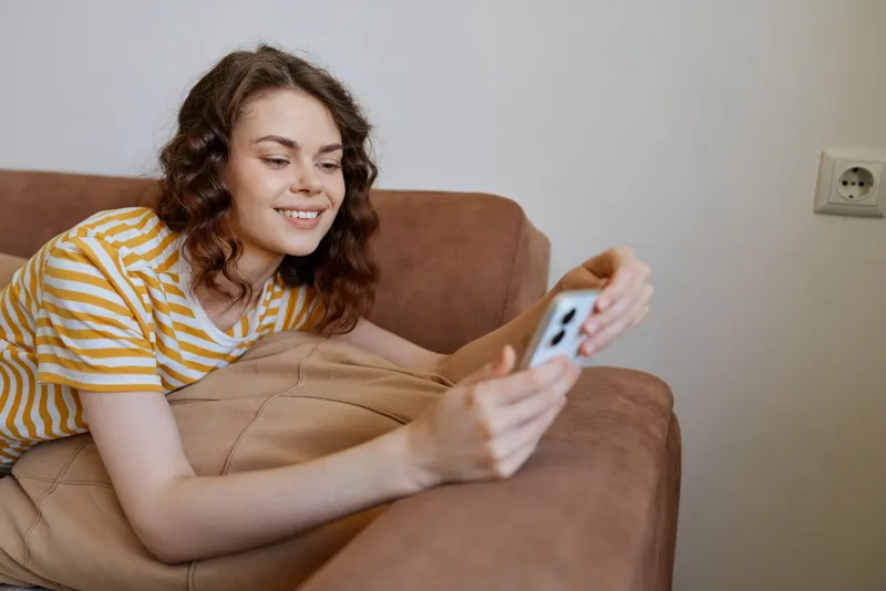 A smiling woman in a striped shirt relaxes on a couch at home, holding a smartphone. A cozy lifestyle moment highlighting comfort, connection, and everyday technology in a bright room.