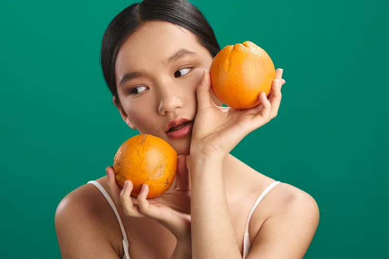 Asian woman holds two oranges near her face in a bright studio with a bold green background, wearing a simple top. She looks calm, conveying freshness and vibrant energy.