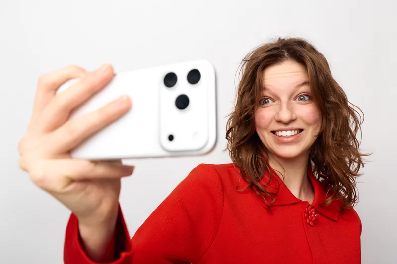 Smiling woman with curly hair wearing red sweater takes selfie with white smartphone on white background. Concept of technology, social media, communication, and casual lifestyle.