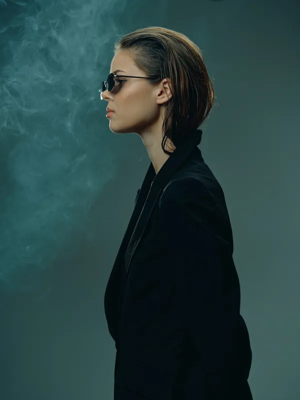Profile of a stylish woman wearing sunglasses and a black jacket with smoky background and moody lighting.