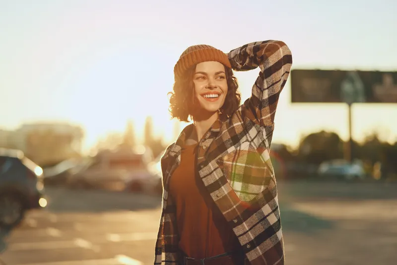 Woman smile sunlight beanie flannel urban portrait candid: Young woman in beanie and flannel smiling in golden hour glow, authenticity and mindful living captured with warm emotional storytelling.