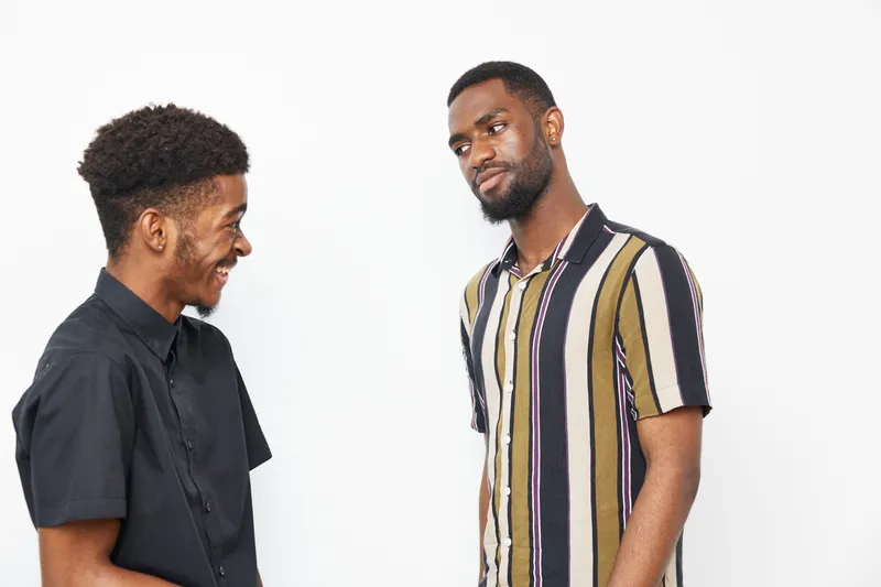 Two smiling African American men share a joyful moment, displaying friendship and camaraderie on a simple white background, with stylish outfits and playful expressions.