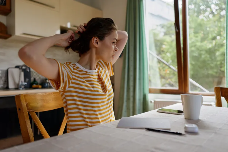 Young woman in a striped t shirt sits at a bright kitchen table, tying her hair back as she prepares for a calm, everyday morning at home, conveying a warm lifestyle moment.